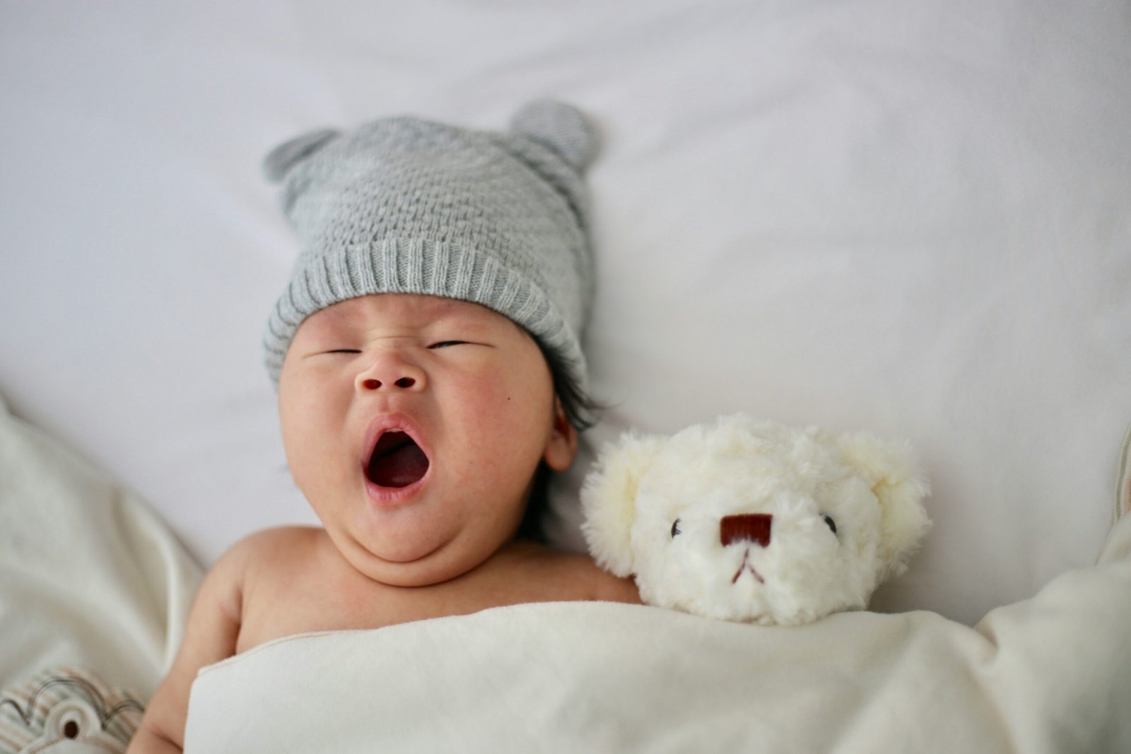 Newborn in a gray knit bear-ear hat yawning beside a small white teddy bear